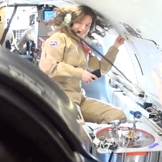 Alessondra Springmann or Sondy, a white person with brown hair wearing a headset and a beige flight suit, floats in reduced gravity inside the cabin of an aircraft. Long brown curly hair extends out from their head. The image quality is a grainy video still from a GoPro camera.